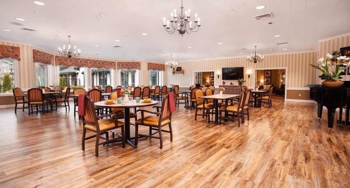 Spacious dining room with multiple tables and chairs arranged neatly. The room features large windows with floral valances, wooden flooring, chandeliers hanging from the ceiling, and a piano with a flower arrangement on top. There is a television mounted on the far wall and warm striped wallpaper.