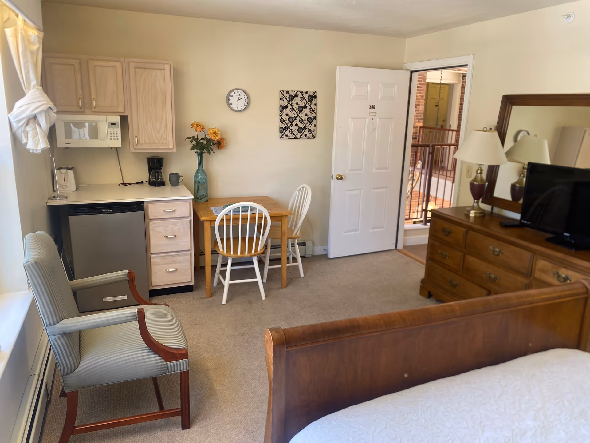 Interior view of a senior living facility room featuring a small kitchenette with a microwave, coffee maker, and dishwasher. There is a wooden table with two chairs, a cushioned armchair near a window with white curtains, a dresser with a lamp and a TV, and a bed partially visible in the foreground. The door to the room is open, showing a balcony and hallway outside.
