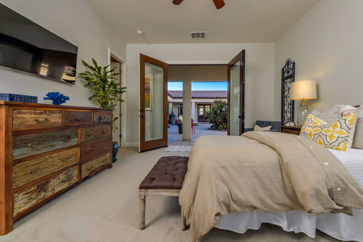 A cozy bedroom with a large bed covered in beige bedding and multiple decorative pillows. There is a wooden bench at the foot of the bed, a wooden dresser with a flat-screen TV mounted above it, and a potted plant beside the dresser. French doors open to an outdoor courtyard with visible plants and buildings under a clear blue sky.
