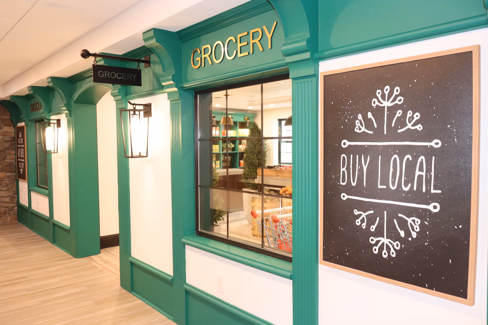 Indoor hallway featuring a green-framed mock grocery storefront with a window and a 'BUY LOCAL' chalkboard sign.