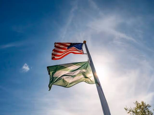 Two flags flying on a flagpole against a blue sky with some clouds. The top flag is the United States flag, and the bottom flag is green and white with a design. The sun is shining behind the flags, creating a bright glow.