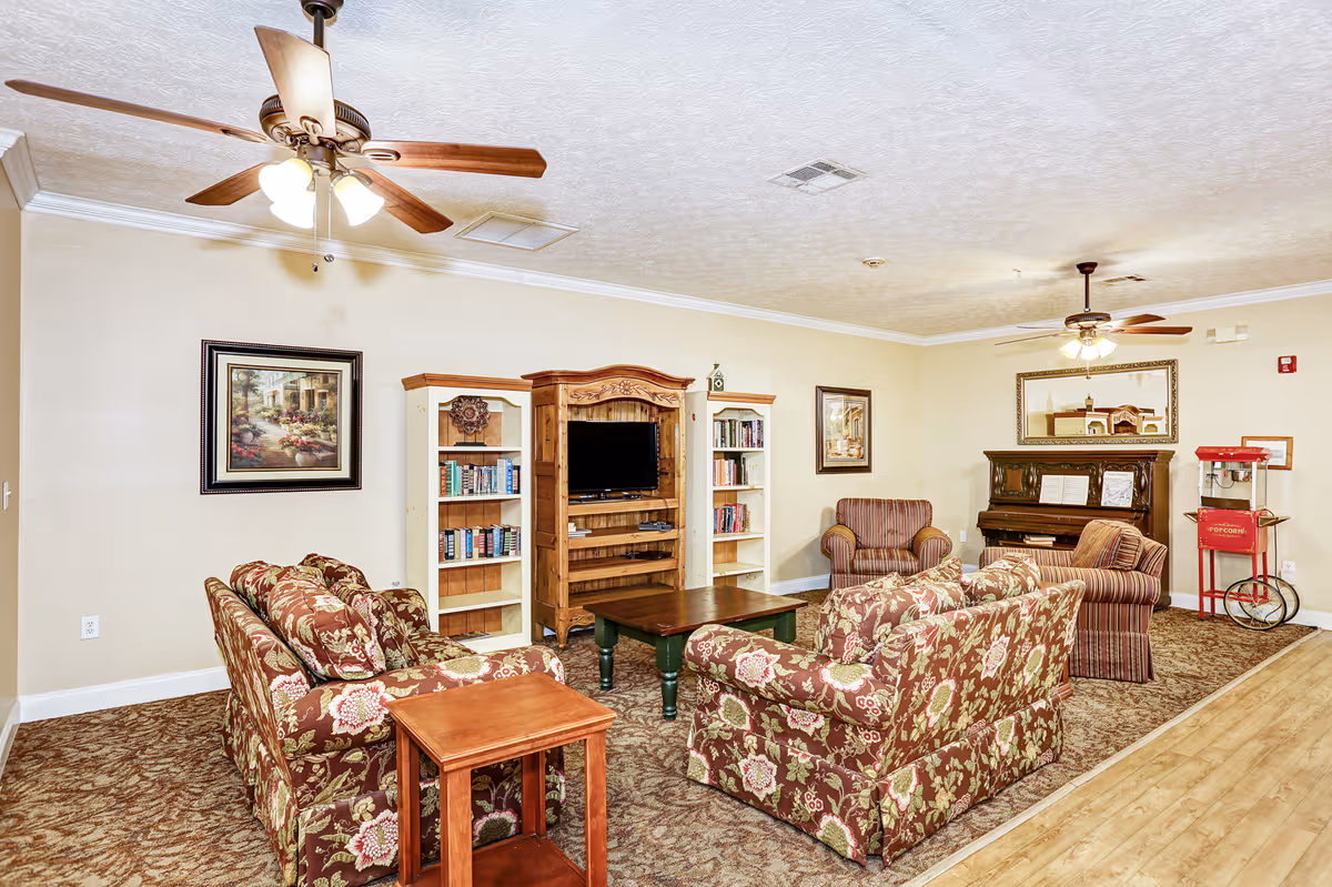 A cozy living room with floral patterned sofas and armchairs arranged around a wooden coffee table. There is a wooden entertainment center with a TV and bookshelves on either side. The room also features a piano with sheet music, a red popcorn machine, two ceiling fans with lights, framed paintings on the walls, and a large mirror above the piano. The floor is carpeted with a patterned rug, and there is a wooden side table next to one of the sofas.
