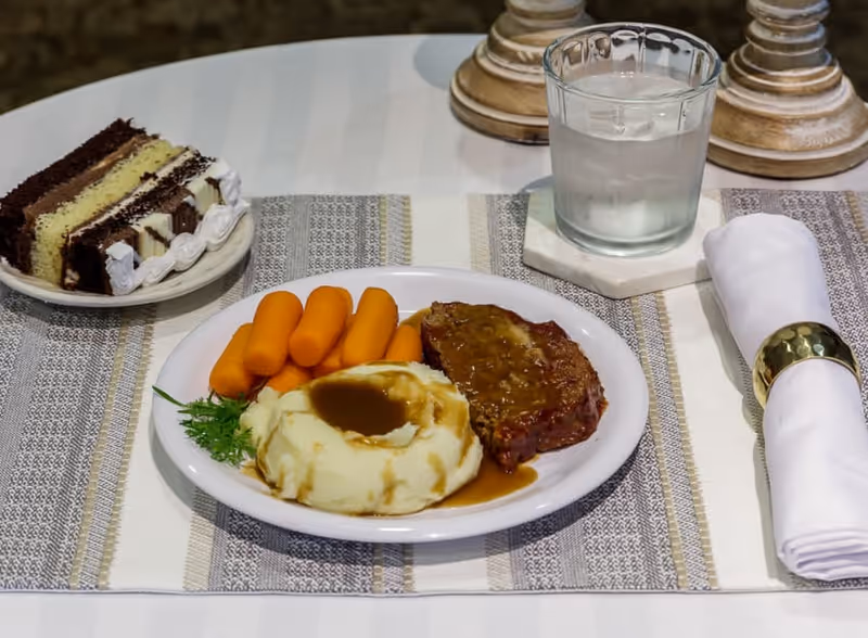 A meal on a white plate consisting of mashed potatoes with brown gravy, cooked carrots, and a piece of meatloaf with gravy. Next to the plate is a glass of water on a coaster and a white cloth napkin with a gold napkin ring. A slice of layered chocolate and vanilla cake with white frosting is on a small plate nearby. The table is covered with a striped placemat.