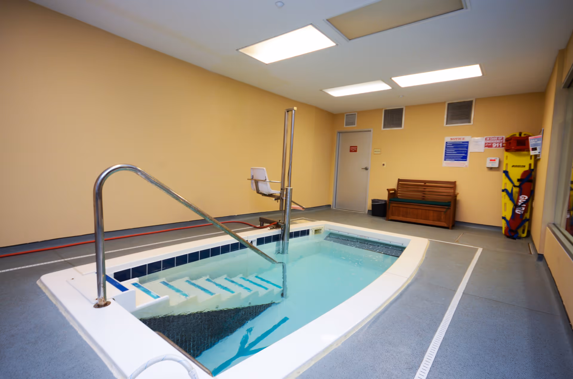 Indoor therapy pool with handrails and a pool lift chair for accessibility, surrounded by beige walls and a gray floor. There is a wooden bench, safety equipment, and emergency signage on the far wall.