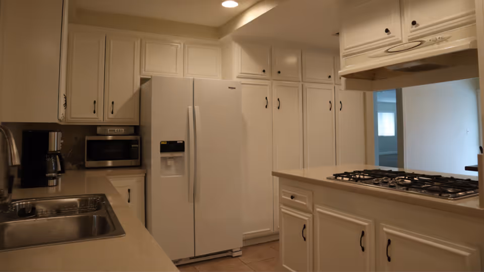 Interior view of a kitchen featuring white cabinets, a double-door refrigerator, a built-in microwave, a coffee maker, a stainless steel sink, and a gas stove on a countertop island. The kitchen has beige countertops and tile flooring, with a doorway leading to another room in the background.