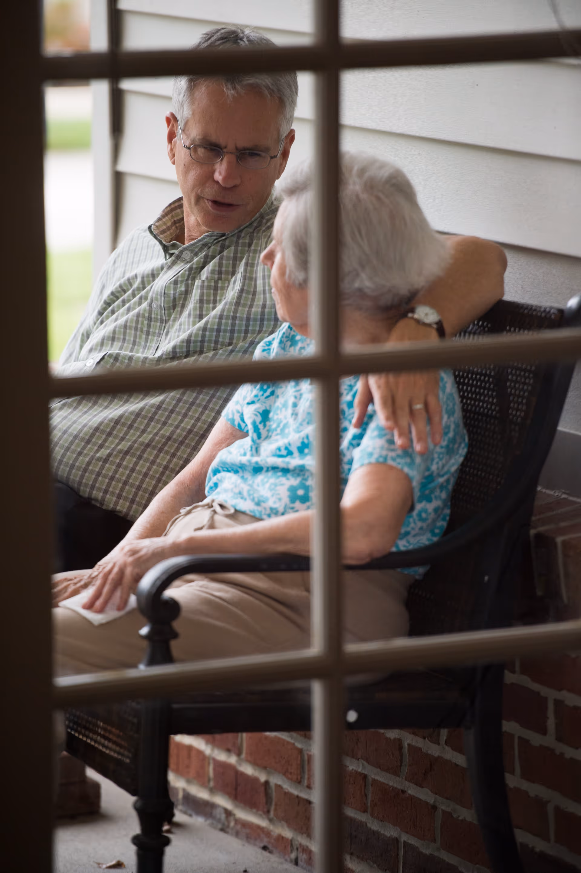 An elderly woman and a middle-aged man sitting closely on a bench outside a building, engaged in a conversation. The man has his arm around the woman, and the scene is viewed through a window with grid-like panes.
