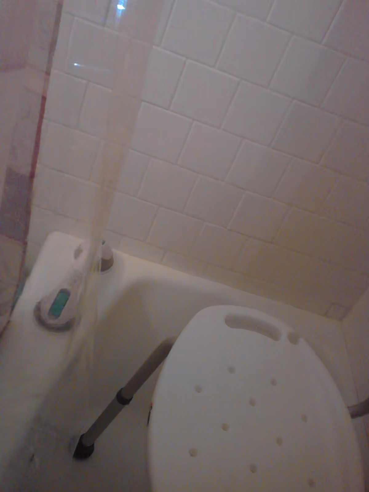 A bathroom shower area with white tiled walls, a white shower chair with holes and metal legs, and a shower curtain partially visible on the left side.