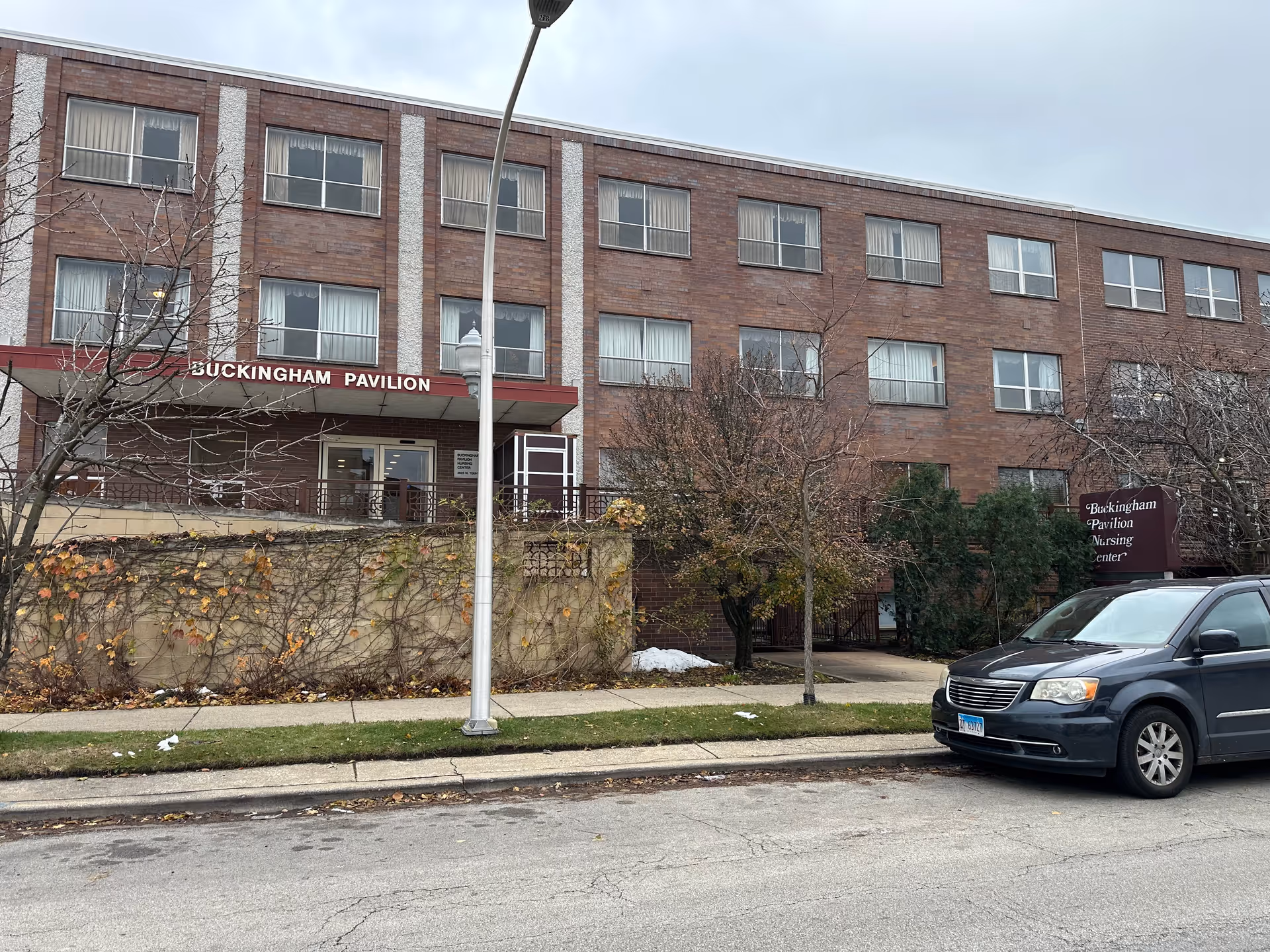 Exterior view of Buckingham Pavilion Nursing and Rehabilitation building, a three-story brick structure with multiple windows and a covered entrance. There is a street lamp and a parked dark-colored minivan in front of the building. Some leafless trees and bushes are visible near the entrance.