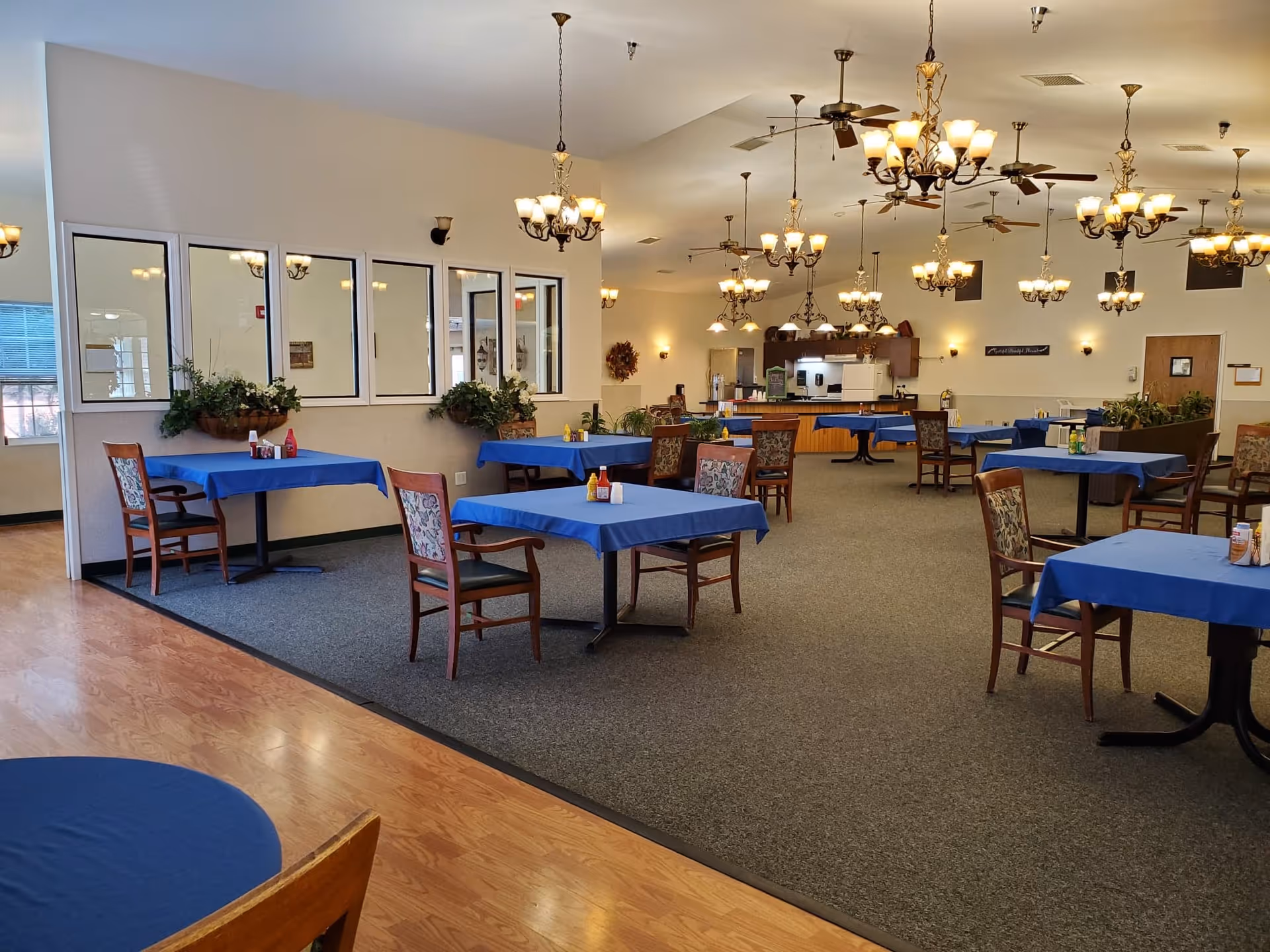 Spacious dining room with multiple tables covered in blue tablecloths, wooden chairs, chandeliers overhead and a service counter in the back.