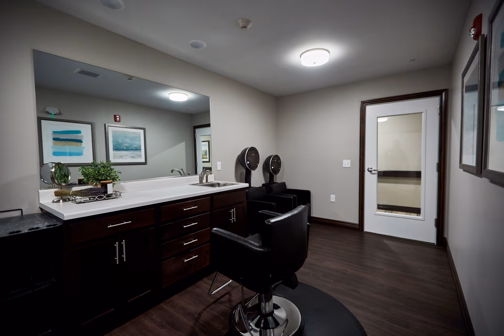 Interior view of a senior living facility's hair salon area with a large mirror above a dark wood cabinet with a white countertop, a sink, two black salon chairs, and two hair dryers mounted on the wall. The room has wood flooring, neutral-colored walls, framed artwork, and a white door with a glass panel.