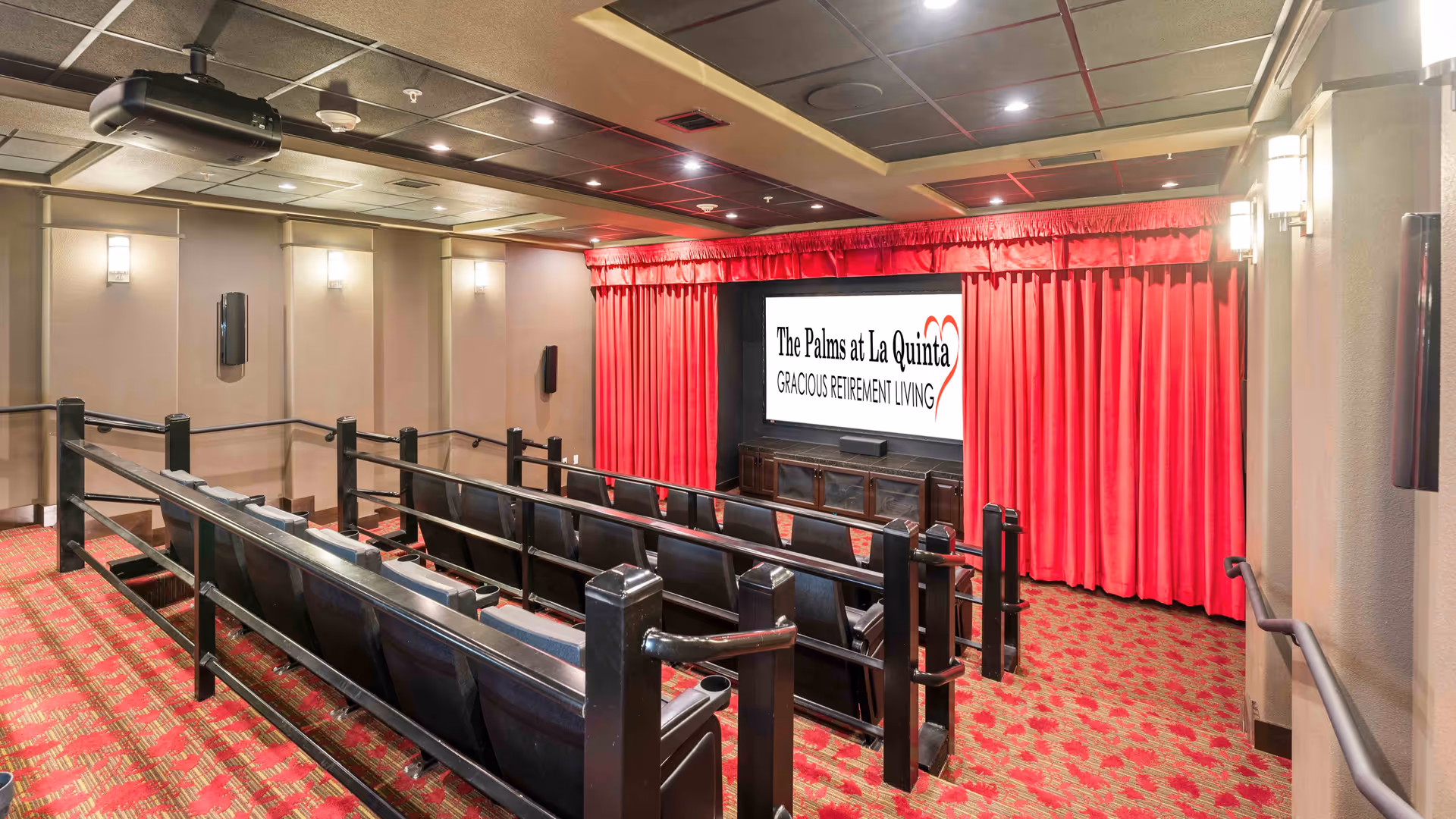 Interior view of a small theater room with tiered seating, black chairs, red patterned carpet, beige walls, and a large screen at the front displaying 'The Palms at La Quinta Gracious Retirement Living' with red curtains on either side.