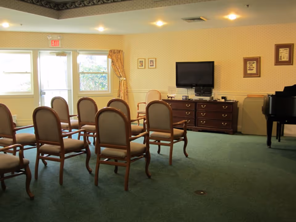 A small meeting or activity room with several wooden chairs arranged in rows facing a flat-screen TV mounted on the wall above a wooden dresser. The room has green carpet, wallpapered walls with framed pictures, a piano on the right side, and windows with yellow curtains letting in natural light.
