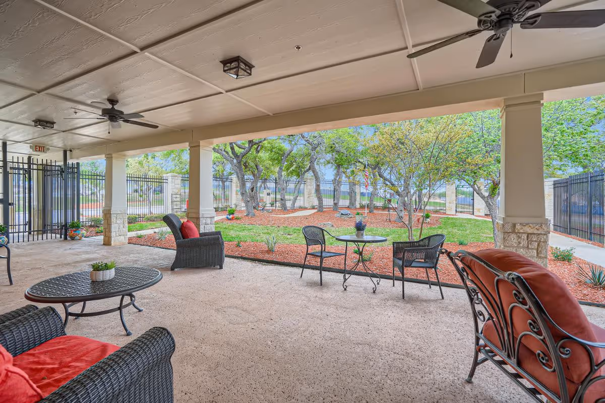 Covered outdoor patio area with ceiling fans and seating including cushioned chairs and small tables, overlooking a landscaped garden with trees and mulch beds enclosed by a black metal fence.