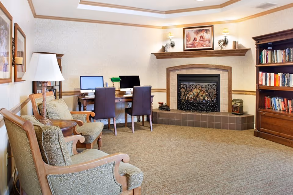 A cozy living room area with two upholstered armchairs and a small round wooden table with a lamp. In the background, there is a wooden desk with two computers and two chairs. A fireplace with a decorative screen is centered on the far wall, above which is a wooden mantel with two wall lamps and a framed painting. To the right, there is a wooden bookshelf filled with books.
