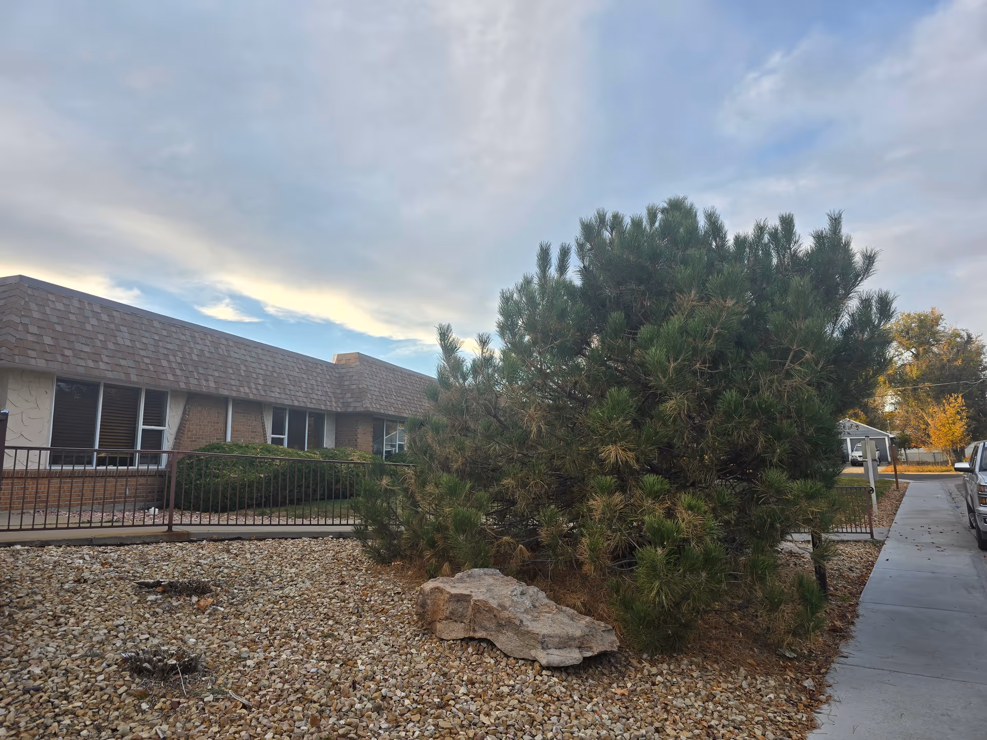Exterior view of Clear Creek Care Center showing a single-story brick building with a shingled roof, a large bush, a rock, and a gravel-covered area in the foreground. A sidewalk runs alongside the building with a parked vehicle visible on the right.