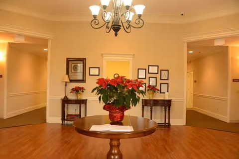Well-lit interior lobby with a central round table holding a red poinsettia, chandelier overhead and framed artwork on the walls.