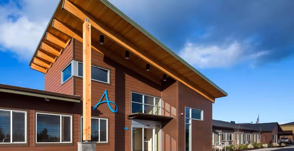 Exterior view of a modern building with brown siding and large windows under a blue sky with some clouds. The building has a prominent wooden beam supporting an extended roof and a large blue letter 'A' mounted on the front wall. There is a pathway and some landscaping visible around the building.