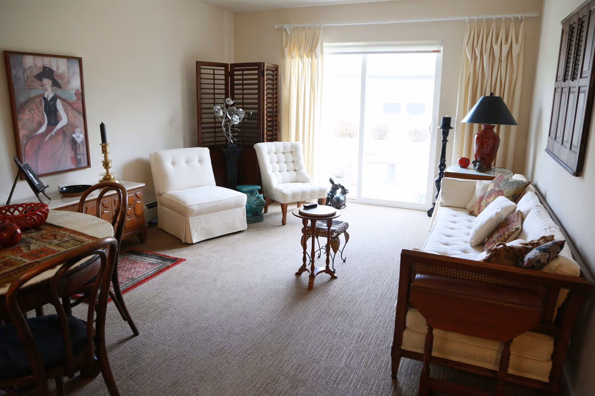 A cozy living room with a beige carpet, two white upholstered chairs, a wooden sofa with white cushions and patterned pillows, a small wooden side table, and a sliding glass door with cream curtains letting in natural light. The room also features a dining table with chairs, a framed painting on the wall, a red lamp on a side table, and decorative items including a black candle holder and a small sculpture.