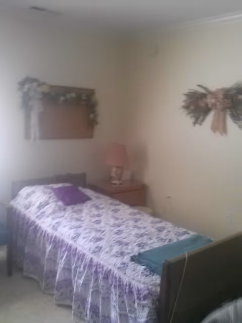 A small bedroom with a single bed covered in a purple floral bedspread and a purple pillow. There is a wooden nightstand next to the bed with a pink lamp on it. The walls are decorated with floral wreaths and a framed piece with flowers.
