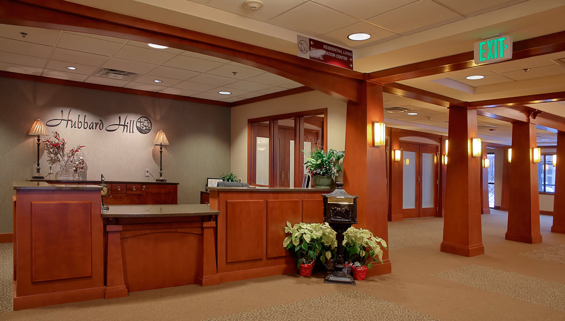 Reception area of Hubbard Hill - Living Wisdom Community featuring a wooden front desk with two lamps, decorative plants, and a sign on the wall reading 'Hubbard Hill'. The space has warm lighting, carpeted floors, wooden columns, and an exit sign above a hallway.