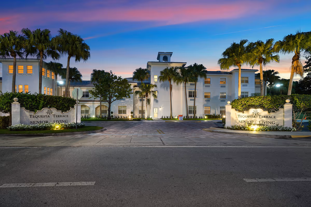 Front entrance of a white senior living building at dusk with palm trees and illuminated signs reading Tequesta Terrace Senior Living.