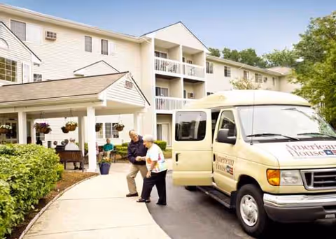 Elderly residents walk toward the entrance of a multi-story senior living building with a parked van.