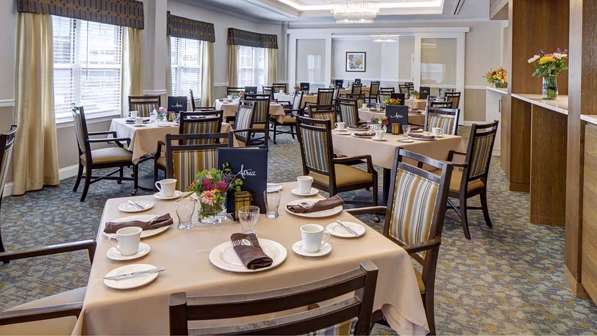 A well-lit dining room with multiple tables covered in beige tablecloths, each set with white plates, cups, glasses, and brown napkins. The chairs have striped upholstery and wooden frames. There are windows with curtains along one wall and a wooden shelving unit with flower arrangements on the right side.