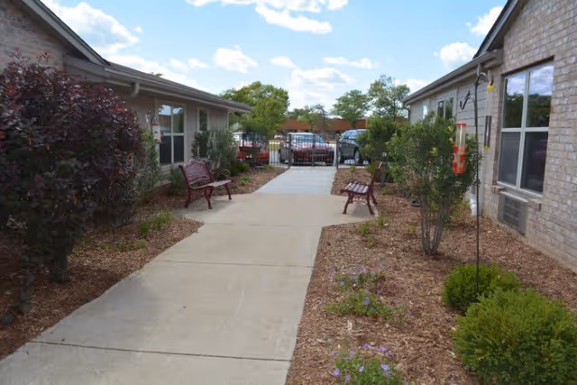 Outdoor walkway between two single-story buildings with benches on either side, landscaped with bushes and small plants, and a clear blue sky with some clouds above.