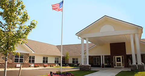 Front entrance of a single-story senior living building with a covered entry, American flag on a flagpole, and landscaped grounds.