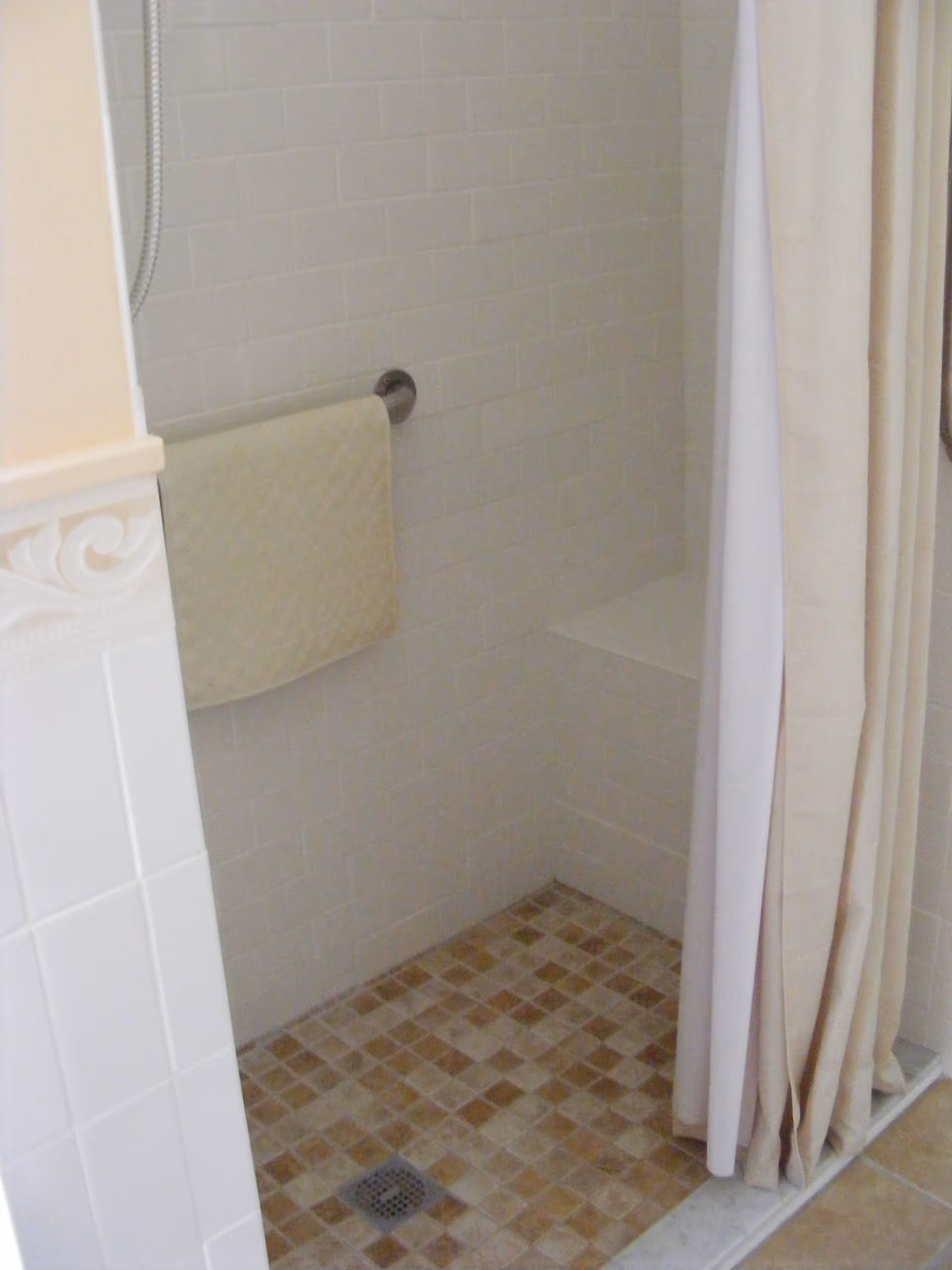 A tiled walk-in shower with a built-in bench, beige and brown mosaic floor tiles, white subway wall tiles, a beige towel hanging on a metal towel bar, and a beige shower curtain partially drawn to the side.