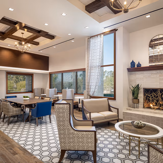 A bright and spacious living room area with a modern fireplace, patterned tile floor, and large windows with blinds and sheer curtains. The room features a mix of upholstered chairs and sofas around a glass coffee table and a wooden dining table with various chairs. The ceiling has wooden beams and recessed lighting.