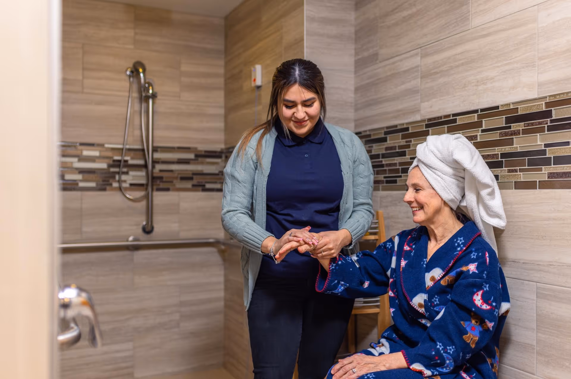 A caregiver assists a seated elderly woman with a towel on her head in a tiled accessible shower room.