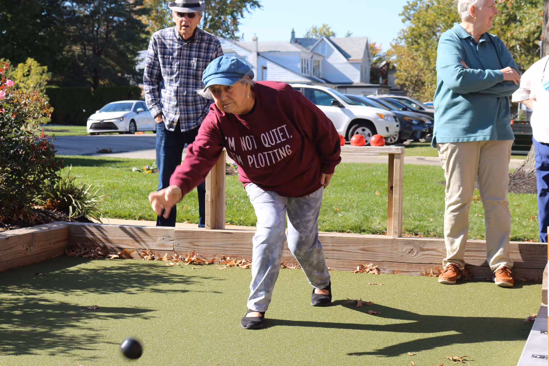 An elderly woman wearing a blue cap and maroon sweatshirt that says 'I'M NOT QUIET. I'M PLOTTING.' is playing bocce ball on a green court outdoors. Behind her, an elderly man in a plaid shirt and hat watches, and two other elderly people stand nearby. There are cars parked and houses in the background under a clear sky.
