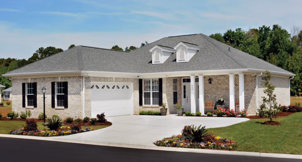 Single-story brick house with a gray shingled roof, white garage door, black window shutters, and a front porch with two chairs. The house is surrounded by a well-maintained lawn, flower beds, and small trees under a clear blue sky.