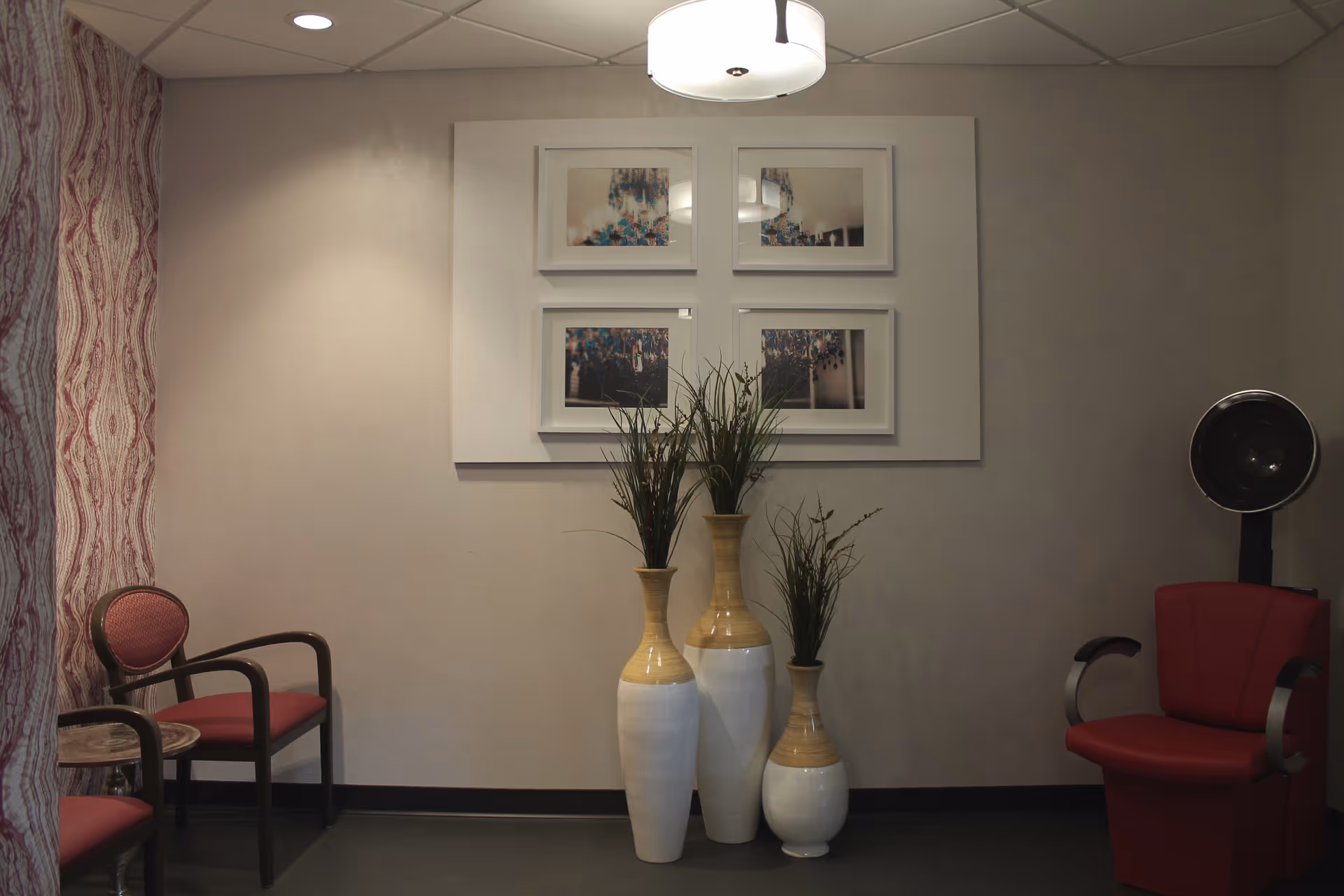 A cozy interior room with two red cushioned chairs, three tall decorative vases with green plants, a wall-mounted set of four framed pictures, and a ceiling light fixture. The wall on the left has patterned wallpaper.