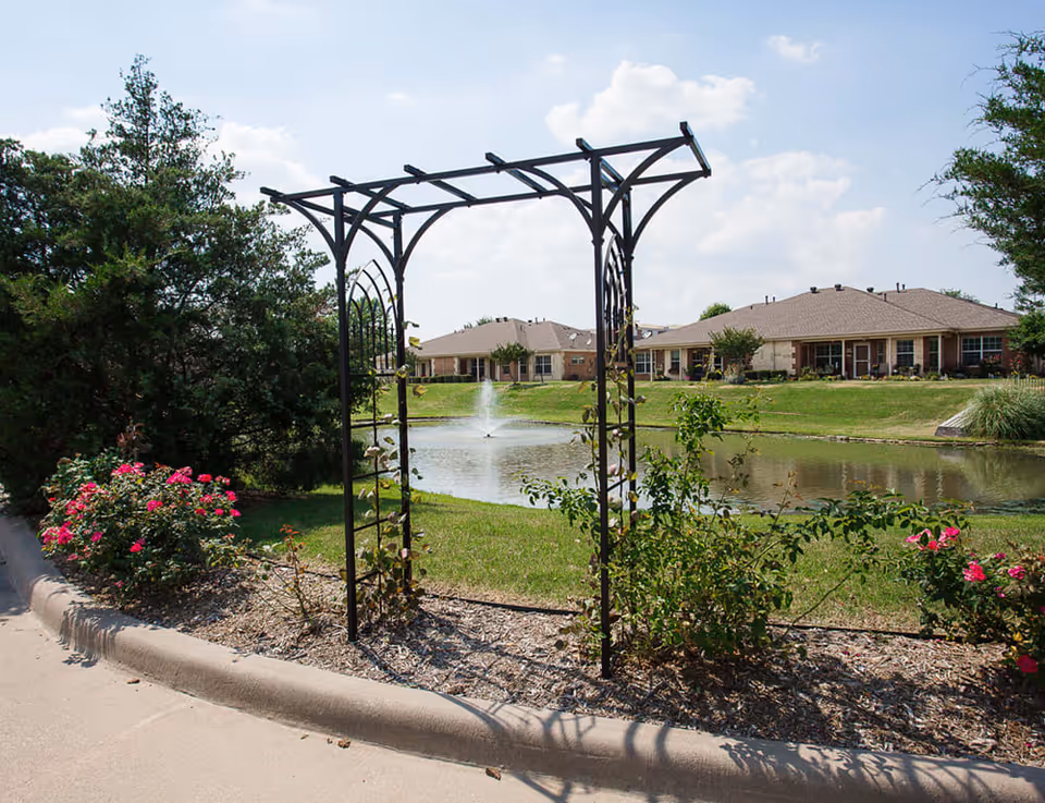 A landscaped outdoor area with a black metal garden arch surrounded by bushes and flowers. In the background, there is a pond with a water fountain and single-story residential buildings under a partly cloudy sky.