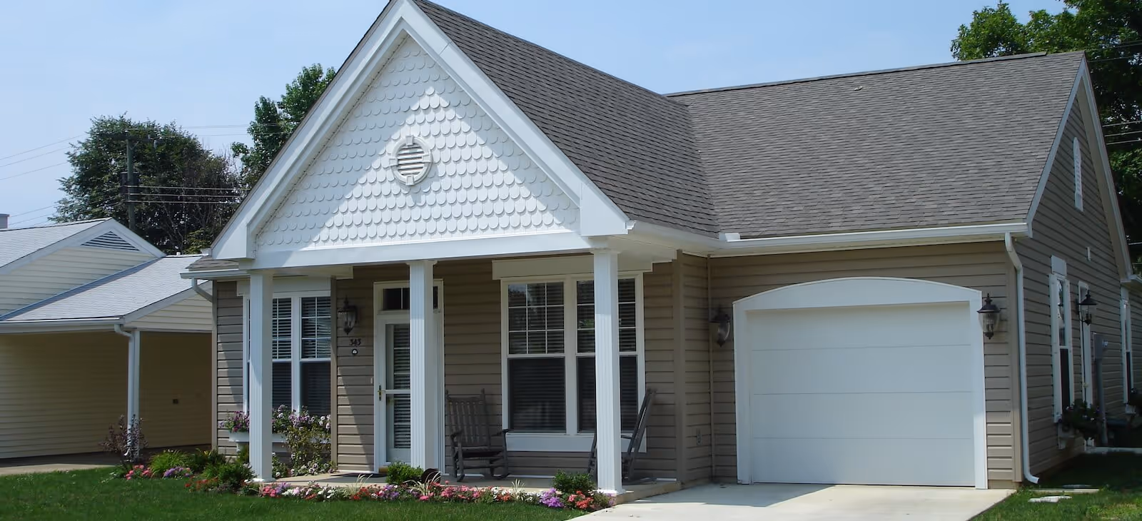 Single-story residential building with beige siding and white trim, featuring a small front porch with two rocking chairs, a white garage door, and a well-maintained lawn with flowers along the front.