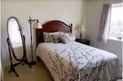 A bedroom with a neatly made bed featuring a white comforter with a brown branch pattern, two pillows, and a wooden headboard. To the left of the bed is a standing oval mirror and a floor lamp. On the right side, there is a nightstand with a lamp and a window with light curtains allowing natural light into the room.