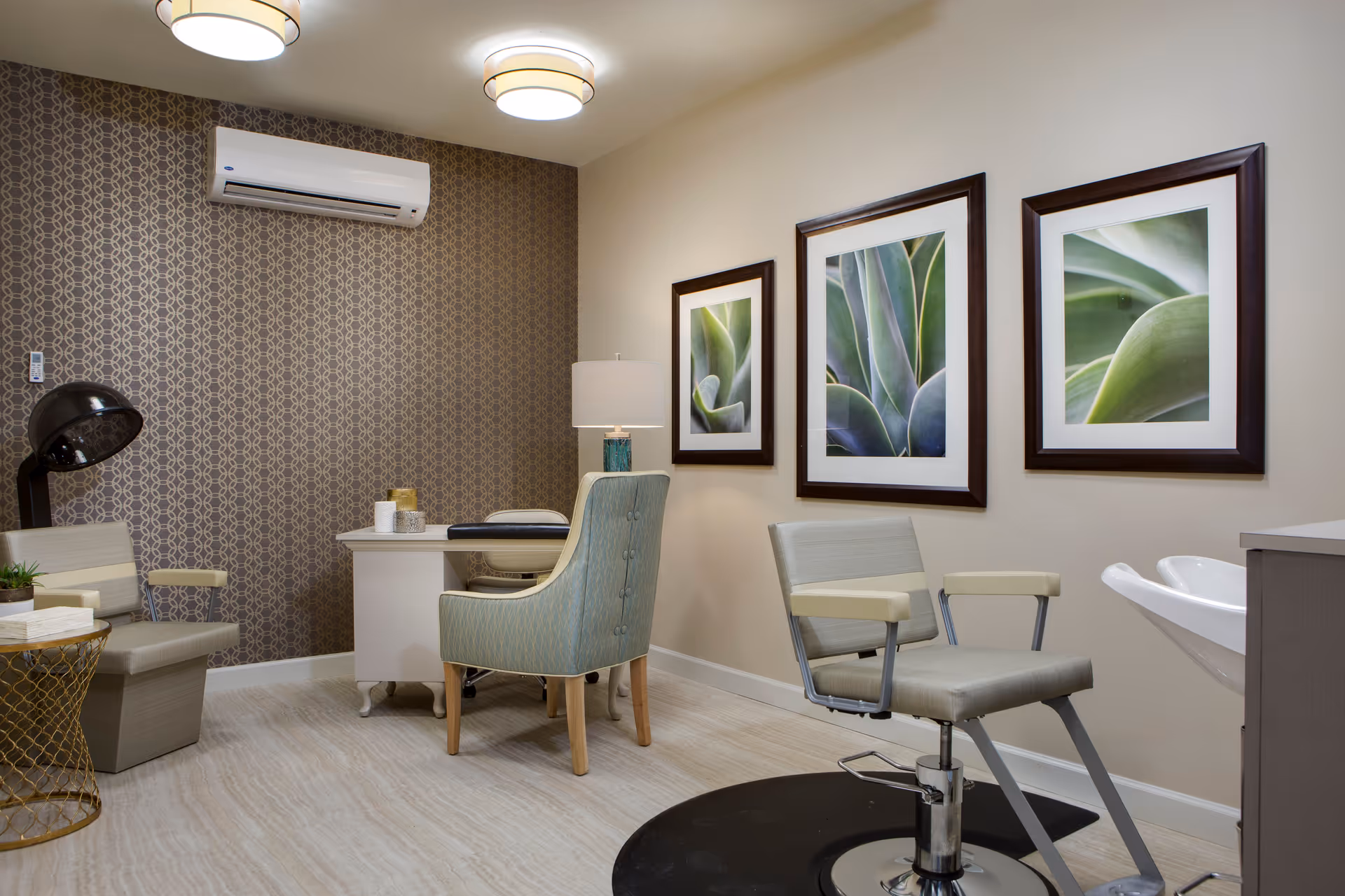 Interior room with a salon chair in front of a white sink, a desk with a blue patterned chair, two beige chairs, a small round gold side table, a black hair dryer, and three framed close-up plant pictures on the wall. The room has a patterned brown accent wall with an air conditioning unit and light-colored flooring.