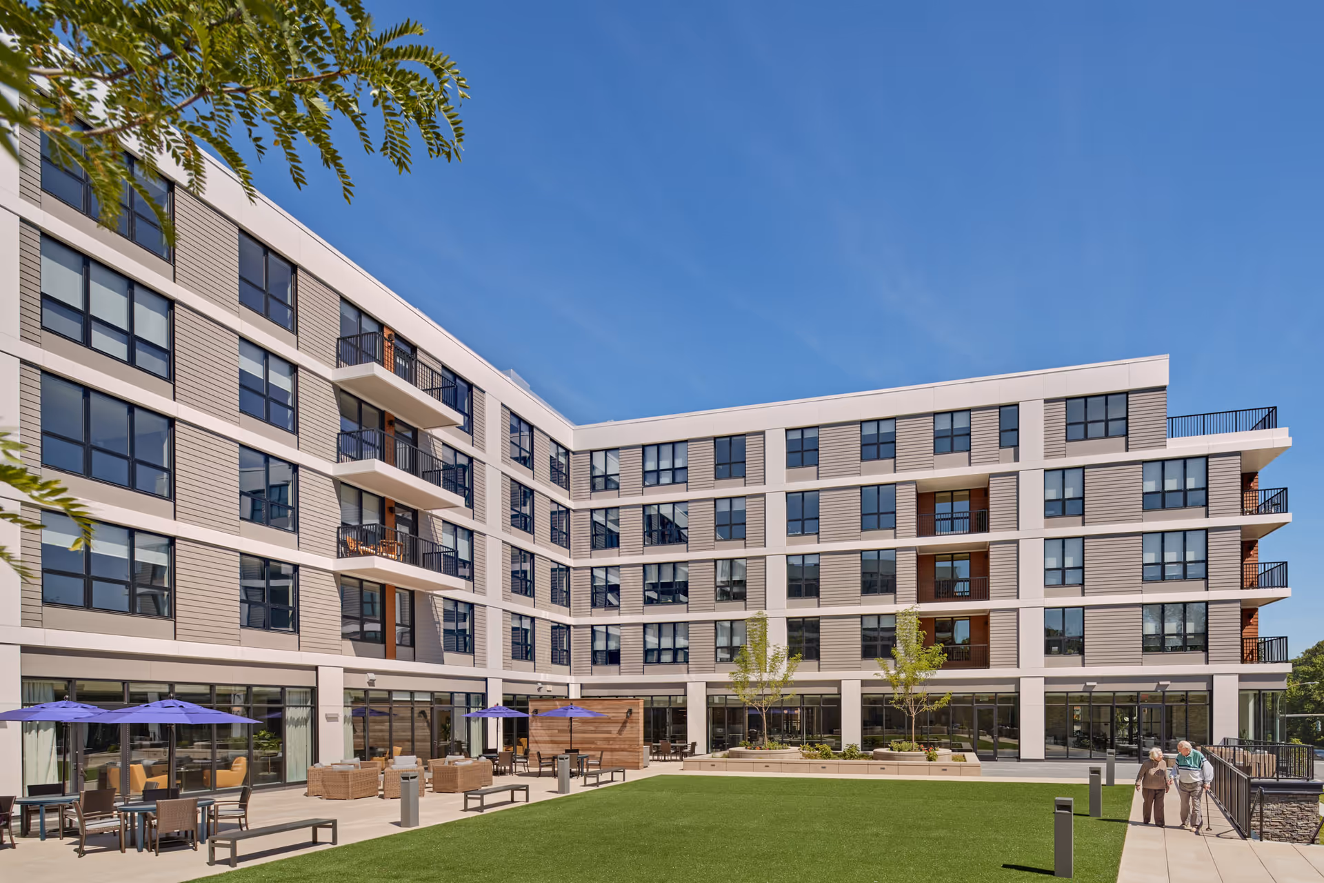 Modern five-story senior living building surrounding a landscaped courtyard with patio seating, umbrellas, and two residents walking on a paved path.