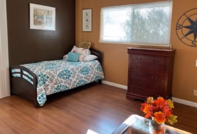 Sunlit bedroom with a single bed, wooden dresser, window, and a small table with a vase of flowers.