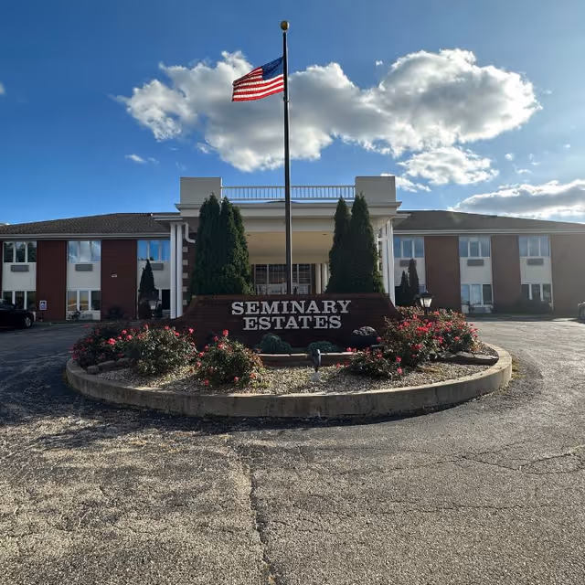 Front exterior view of Seminary Estates building with an American flag on a flagpole in the center of a circular flower bed with bushes and flowers. The building is two stories with multiple windows and a covered entrance.