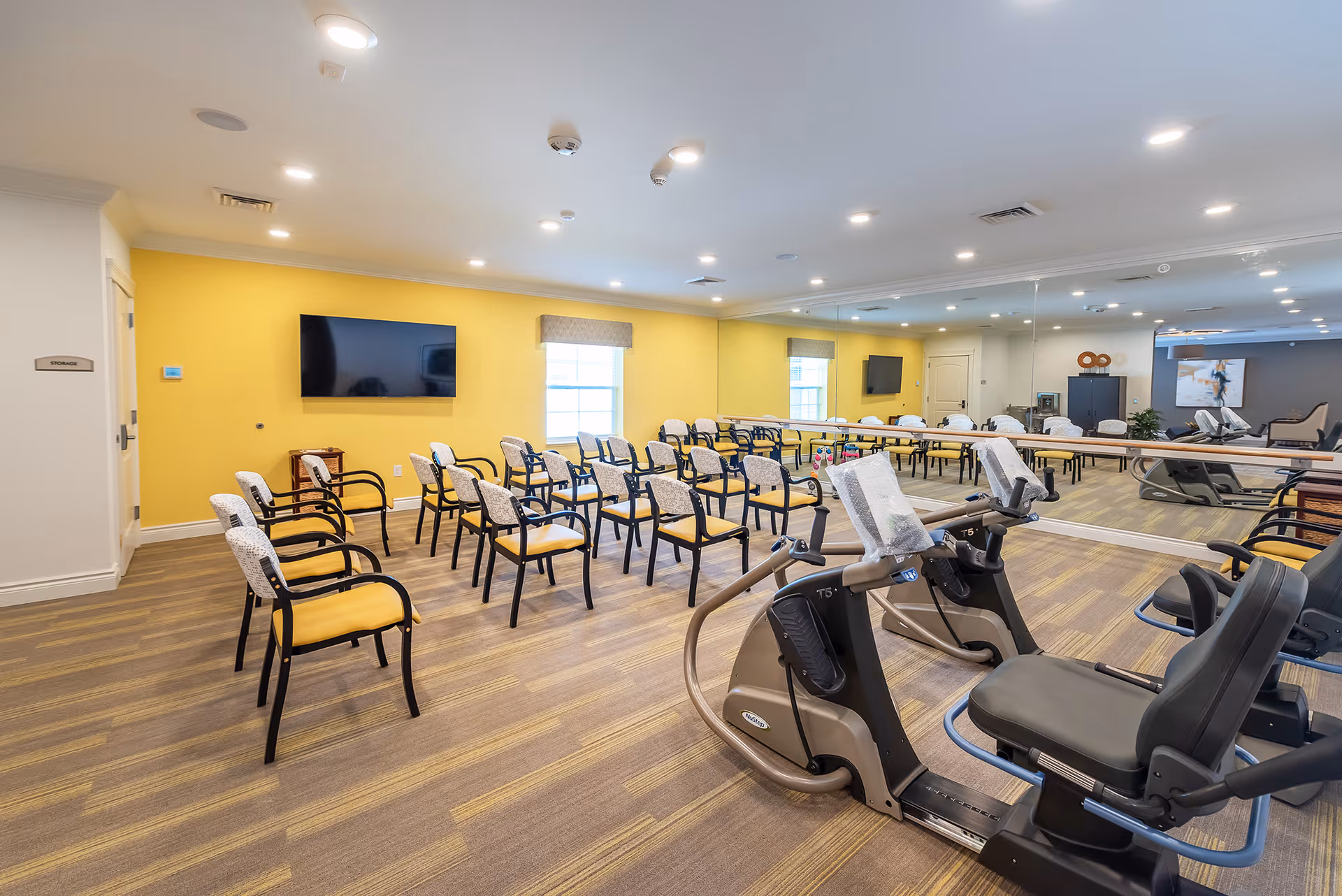 A bright multipurpose activity room with rows of chairs, recumbent exercise bikes, a wall-mounted TV on a yellow accent wall and a large mirrored wall.
