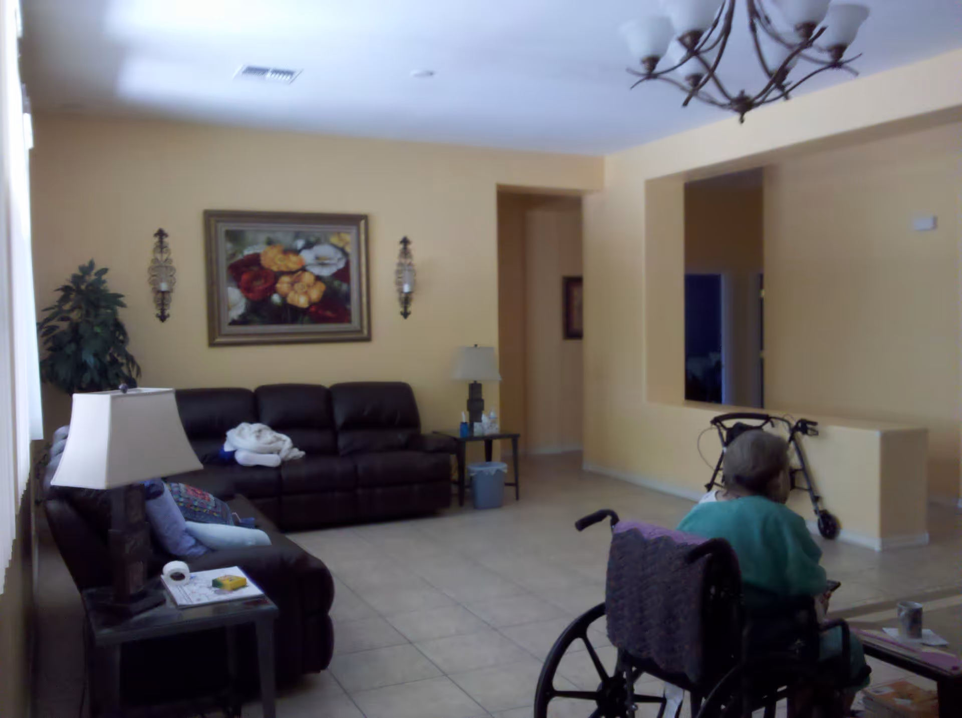 Interior view of a living room in an assisted living facility with beige walls, a dark brown leather sofa, a side table with a lamp, a painting of flowers on the wall, a chandelier on the ceiling, and an elderly person in a wheelchair facing away from the camera. A walker is also visible near a half wall.