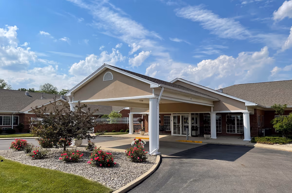 Entrance of Forest Glen Health Campus - Memory Care with a covered drop-off area supported by white columns, surrounded by landscaped flower beds with red flowers and bushes under a partly cloudy blue sky.