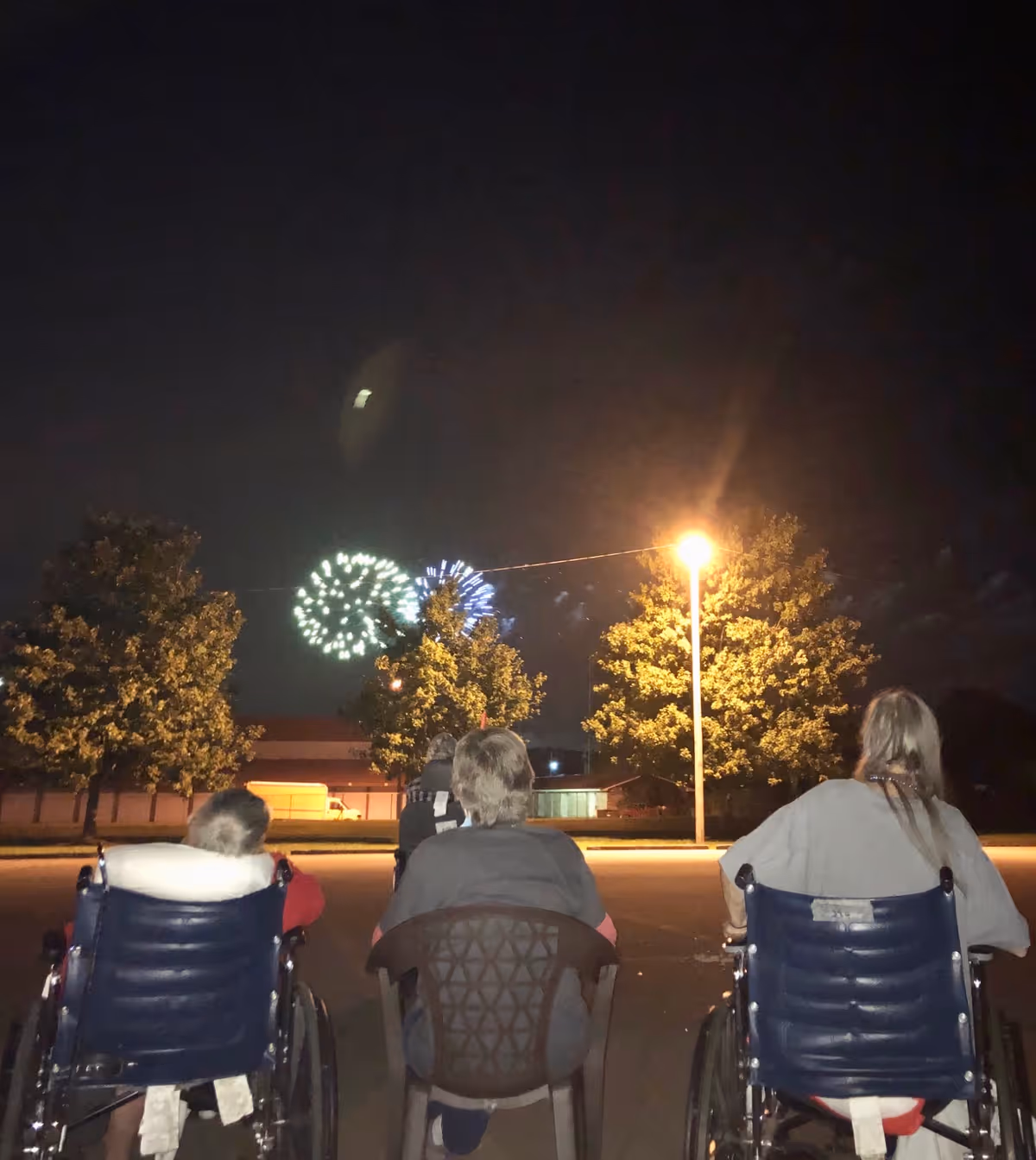 Three elderly individuals, two in wheelchairs and one seated in a plastic chair, watching fireworks in the night sky outdoors near a streetlight and trees.