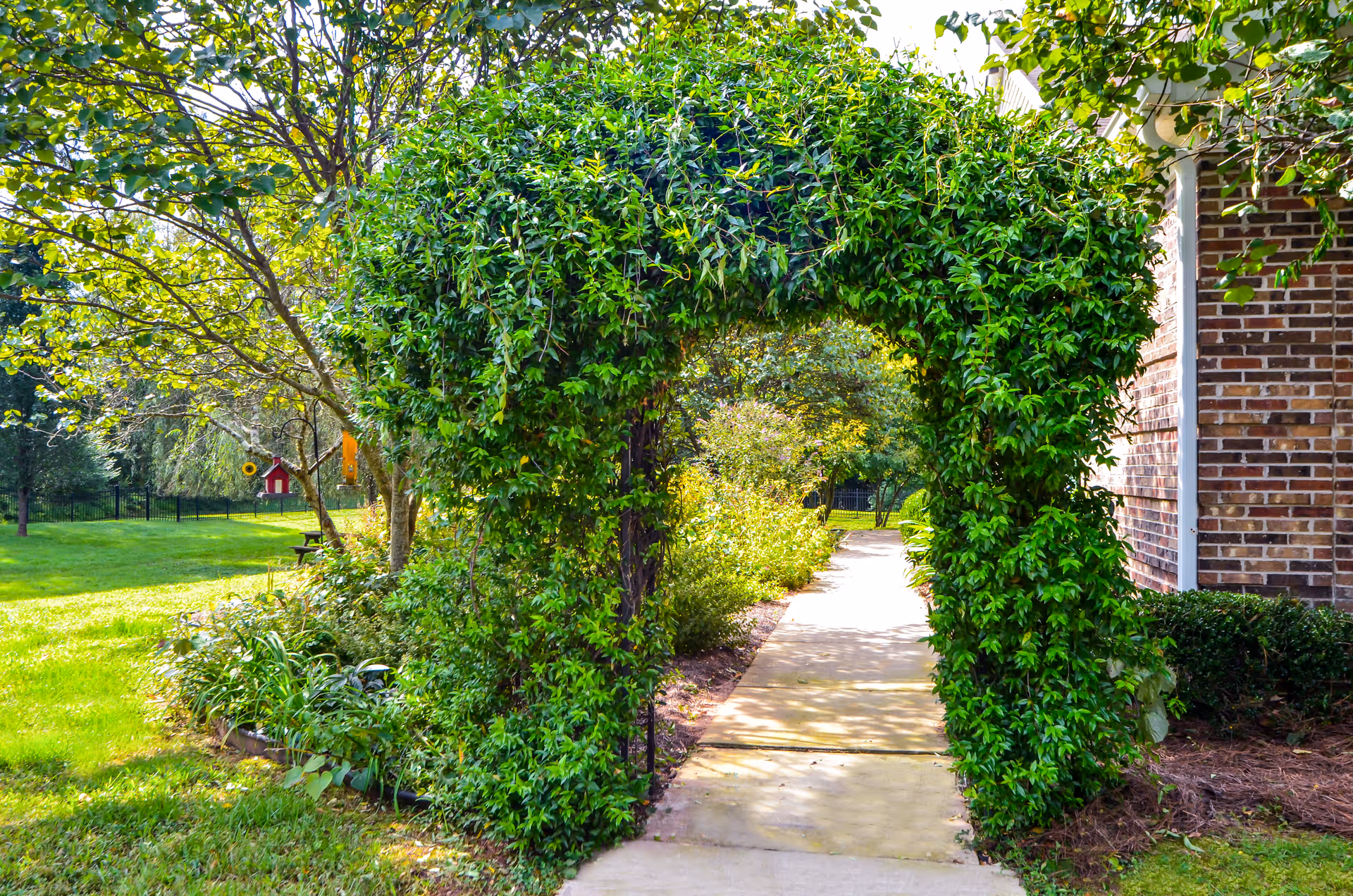 A garden pathway with a lush green archway covered in climbing plants. The path leads past a brick building on the right and is surrounded by grass, trees, and various plants under bright daylight.