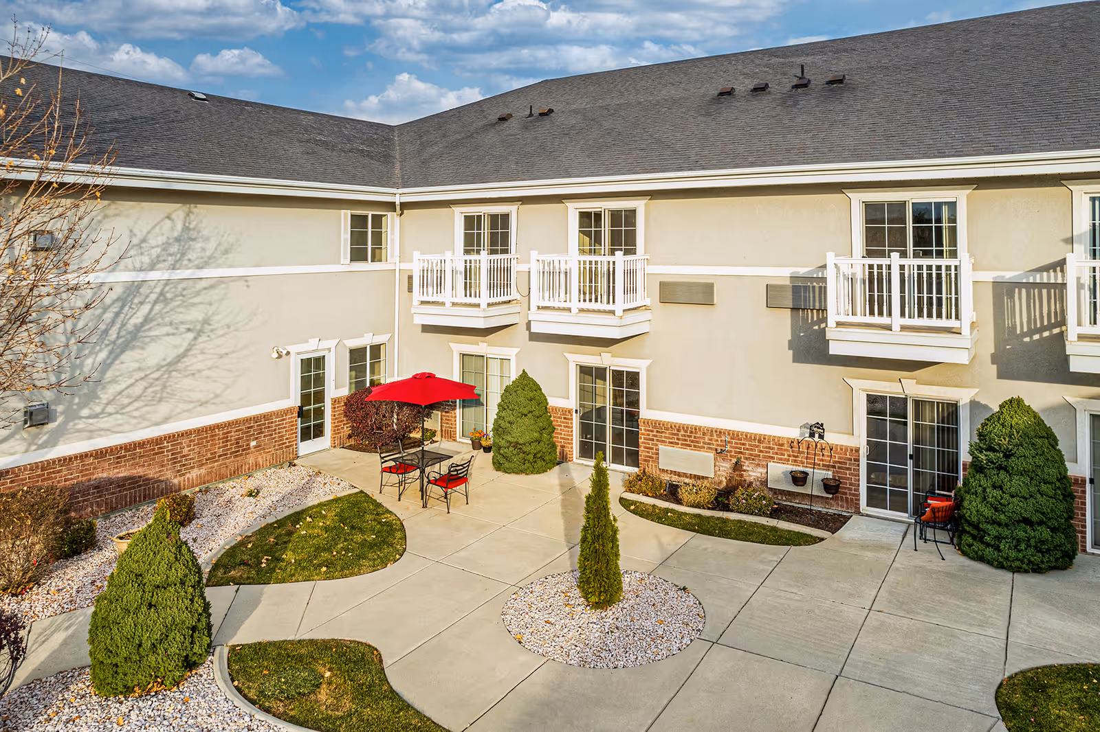 Sunlit courtyard of a senior living building with patio tables and a red umbrella, small balconies, and landscaped walkways.