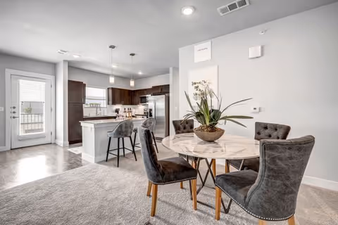 Open-plan interior showing a round dining table with four upholstered chairs next to a kitchen island with pendant lights and stainless-steel appliances.