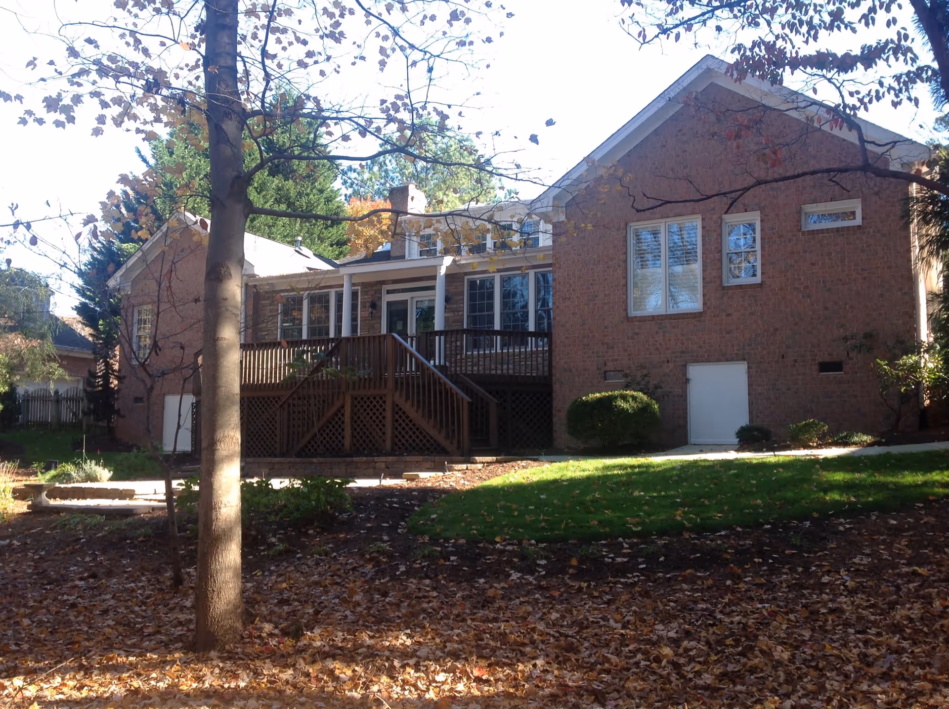 Back view of a brick residential building with a wooden deck and stairs leading down to a yard covered with fallen leaves. Trees with autumn foliage surround the area, and there is a patch of green grass near the building.